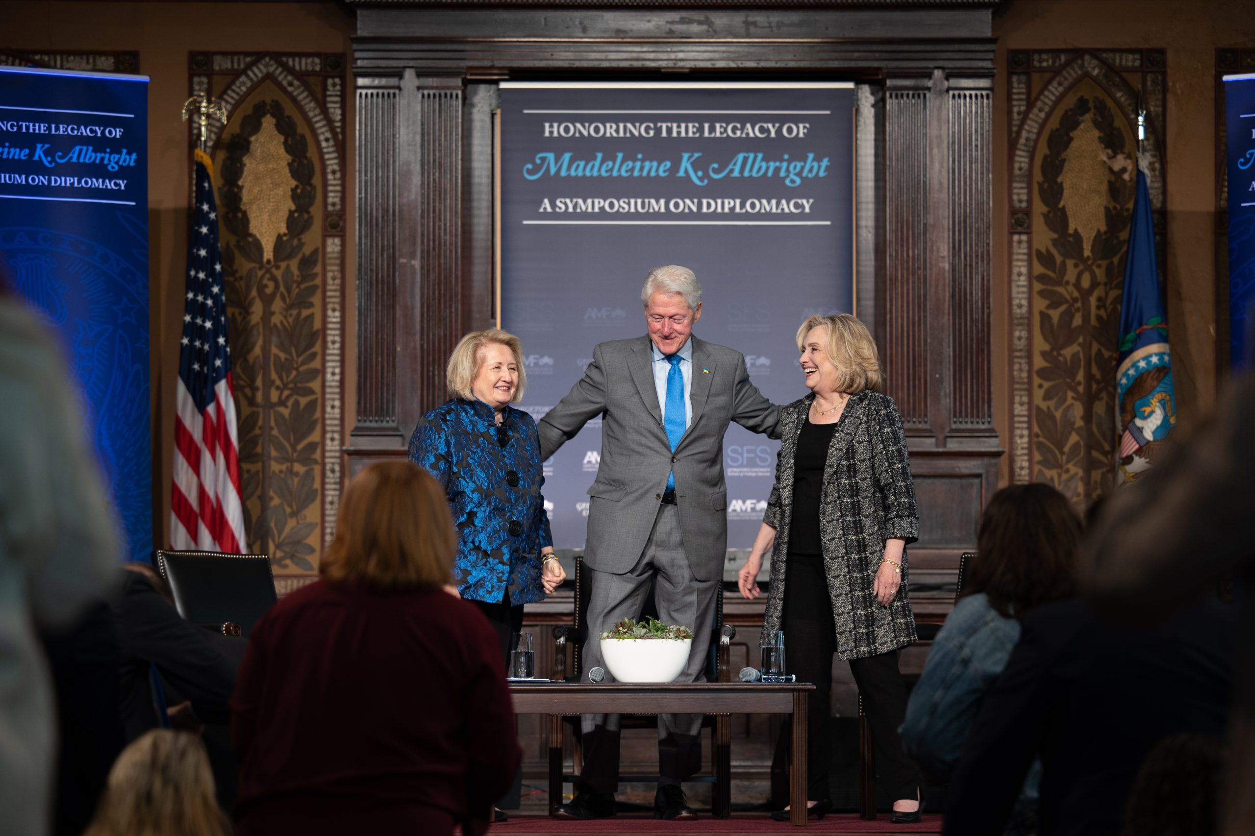 Melanne Verveer, Bill Clinton and Hillary Clinton stand on a stage with a banner behind them with the text "Honoring the Legacy of Madeleine Albright"