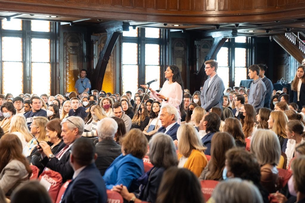 Students stand in line in the aisle of the audience to ask questions at the microphone while one young woman delivers her questions.