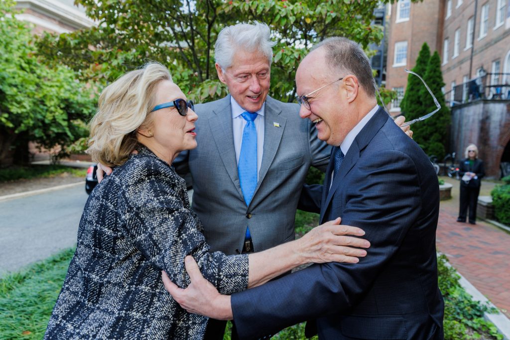 Hillary Clinton wears sunglasses while she embraces President DeGioia and Bill Clinton stands in the middle with his arms around both.
