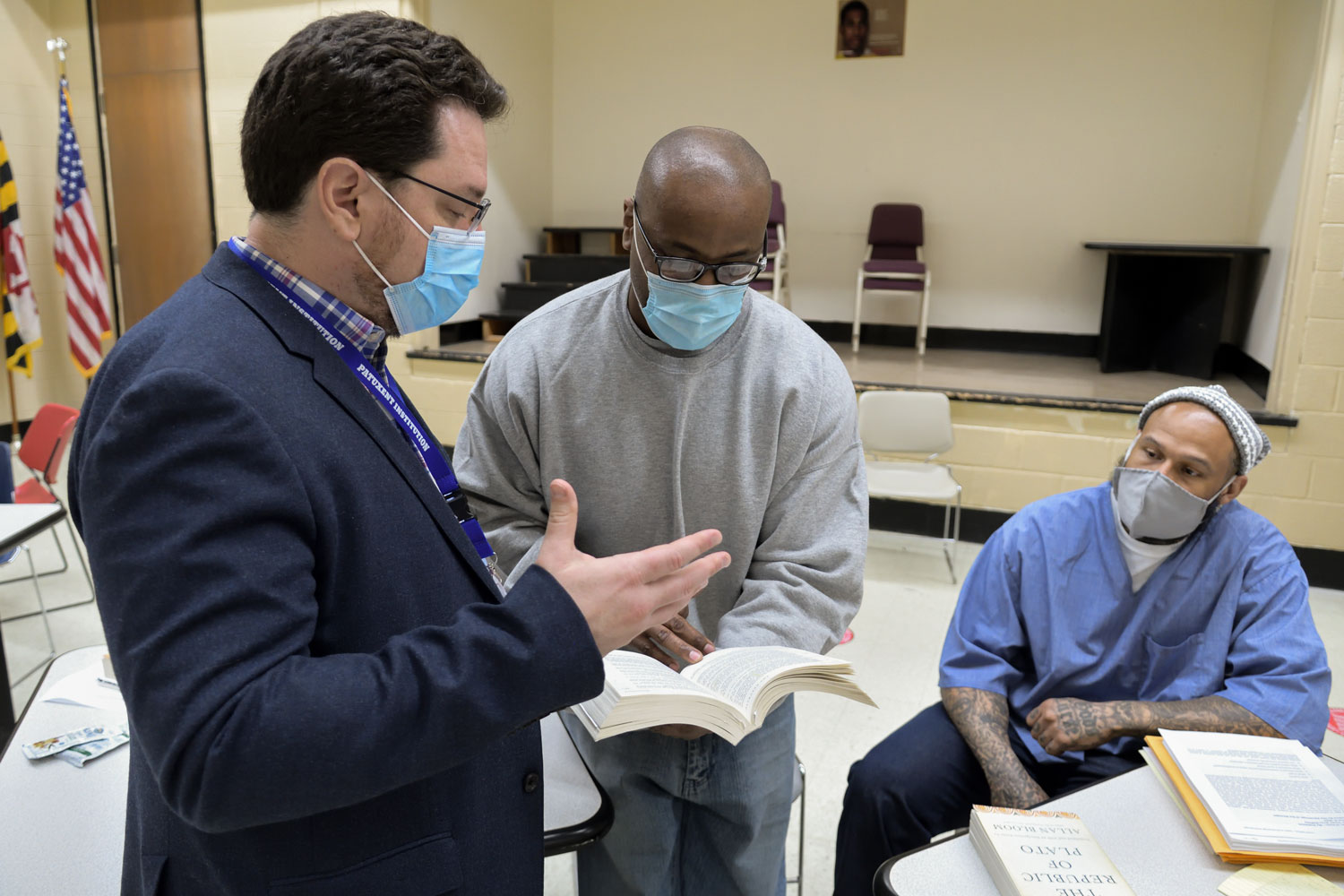 Man wearing a mask holds a book and discusses with two other men wearing masks in a prison