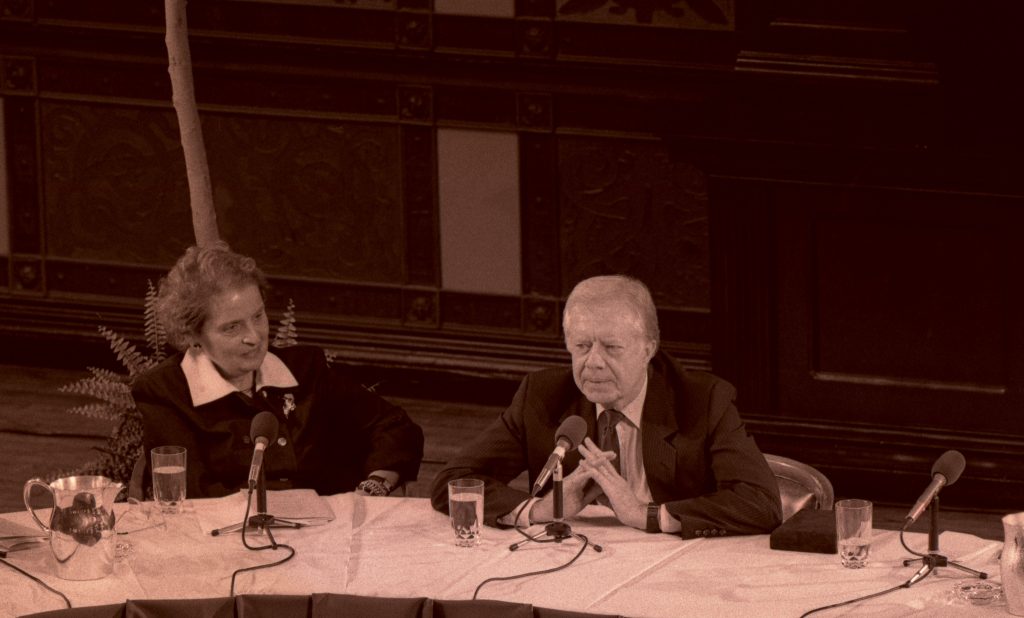 Madeleine Albright with Jimmy Carter in sitting behind microphones on a table