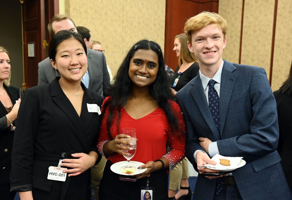 Three students in formal clothes hold plates and smile at the camera