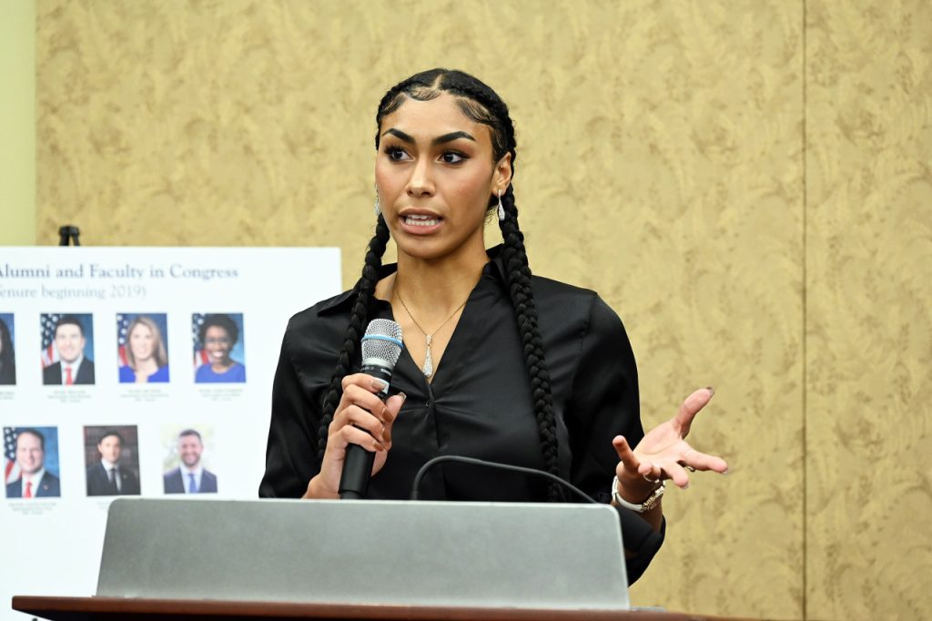 Young woman with two braids and a black blouse speaks into a microphone