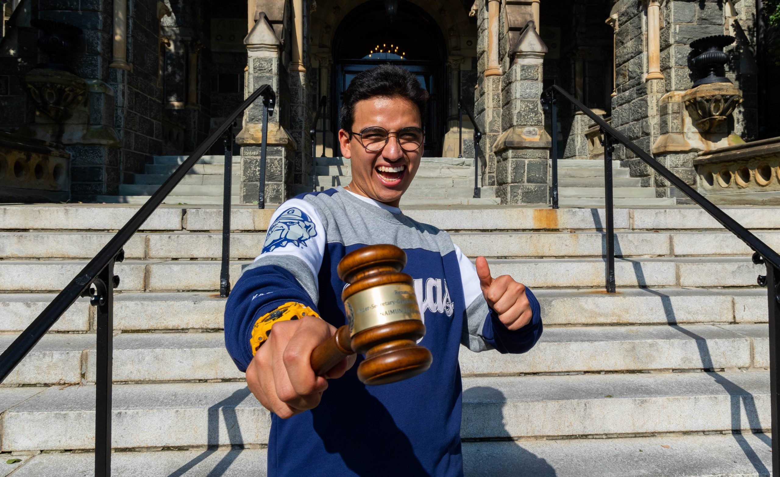 Student in a blue and gray Hoyas sweatshirt holds a gavel and gives a thumbs up