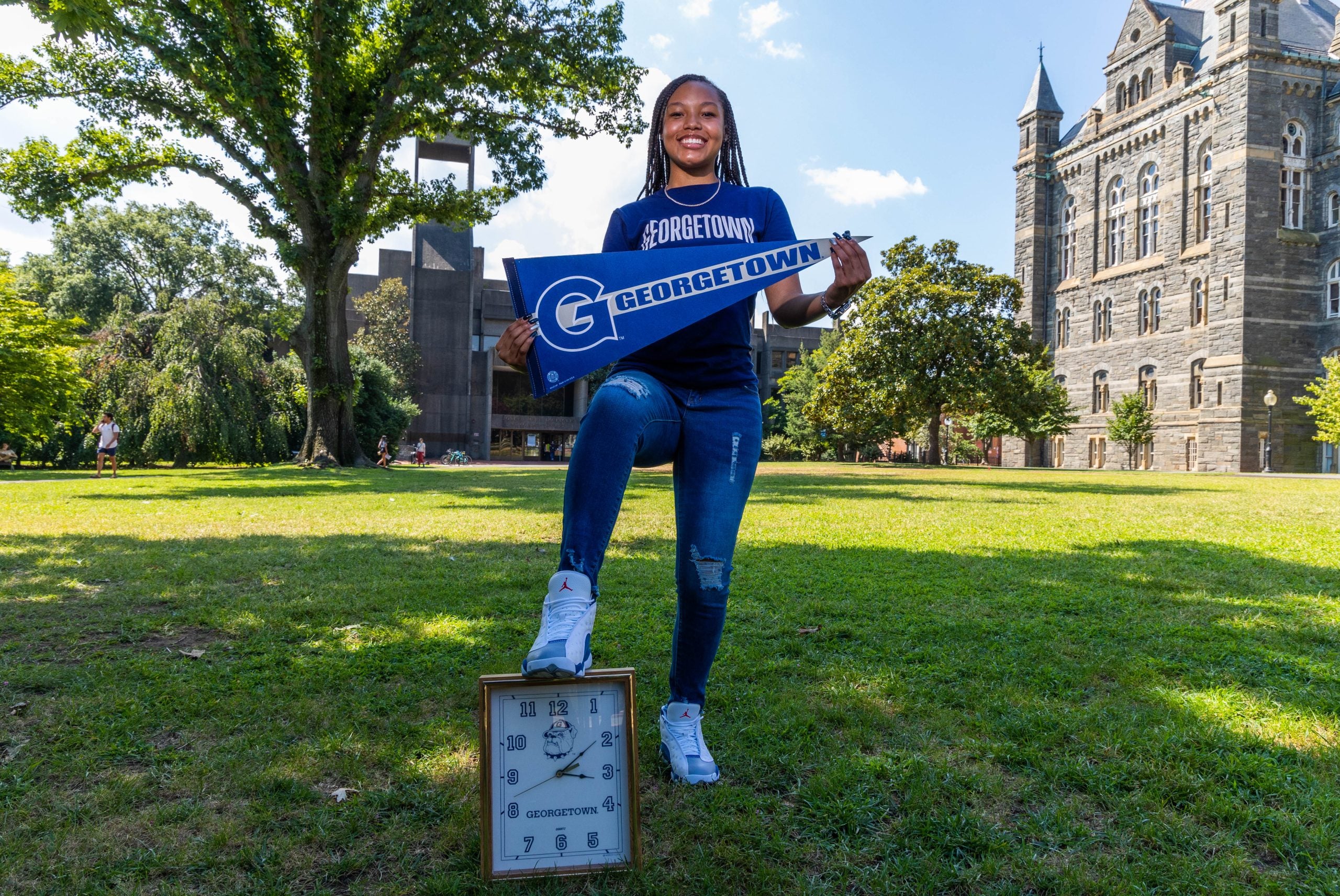 Young woman wearing a blue Georgetown T-shirt holds a blue Georgetown pennant and puts her foot on a small clock in the grass