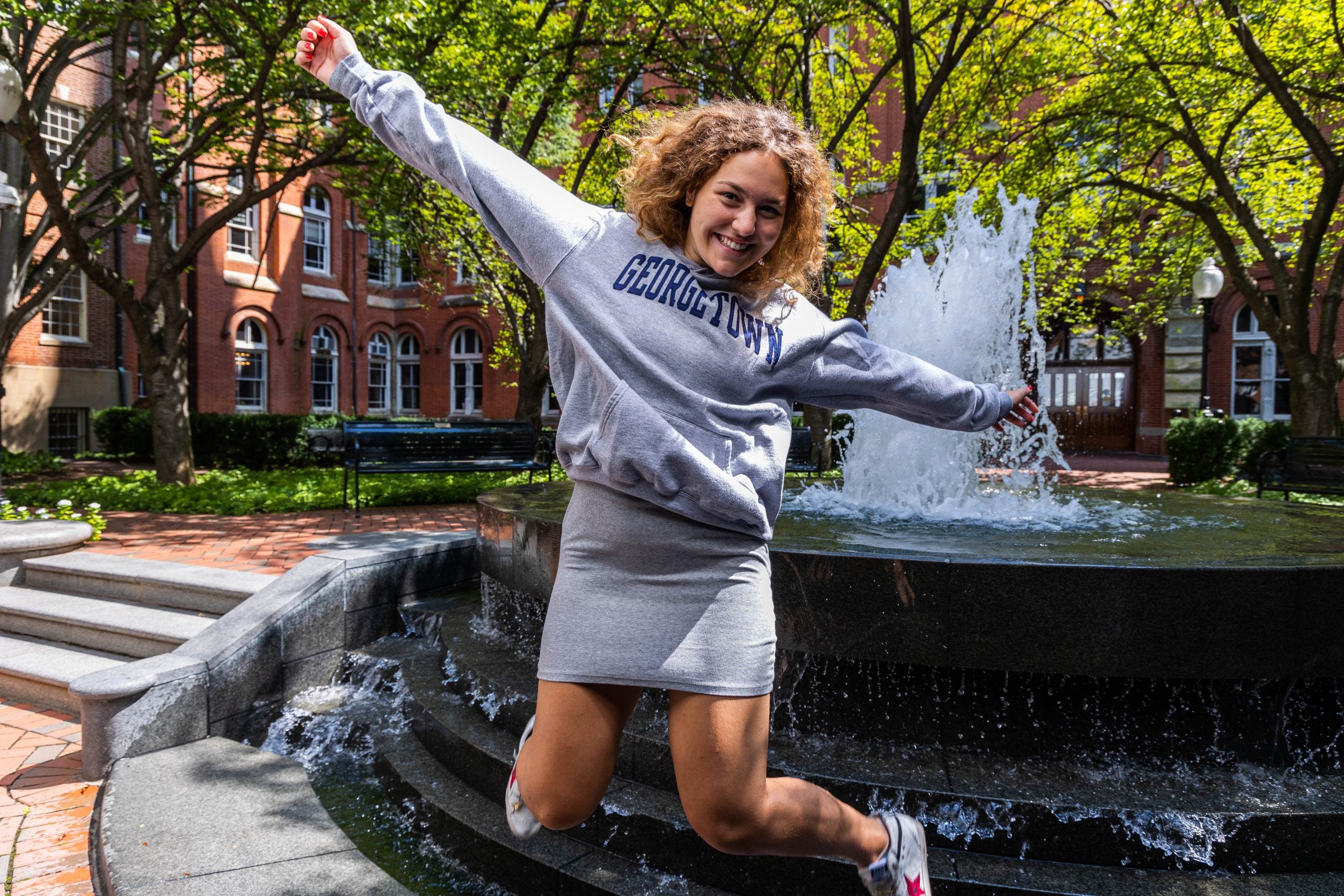 Young woman wearing a gray Georgetown sweatshirt and skirt jumps in front of a fountain