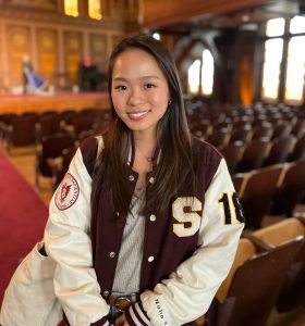 Young woman in a letterman jacket stands in an empty Healy Hall