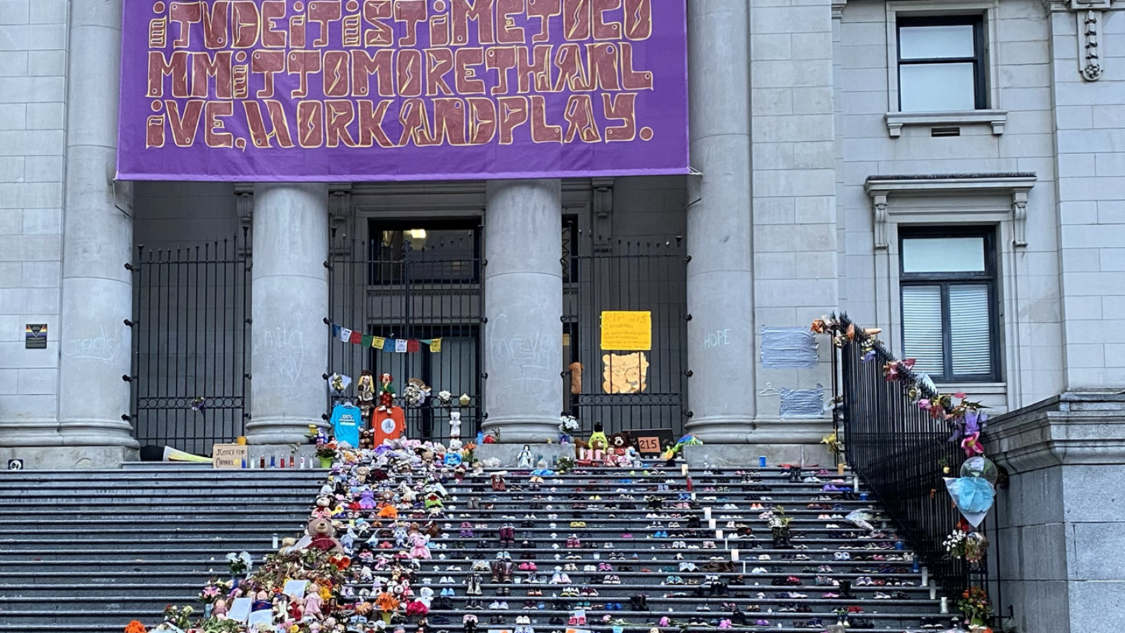 Children&#039;s shoes, flowers, and other symbolic items line the steps of the Vancouver Art Gallery as a purple banner hangs over its stone columns.