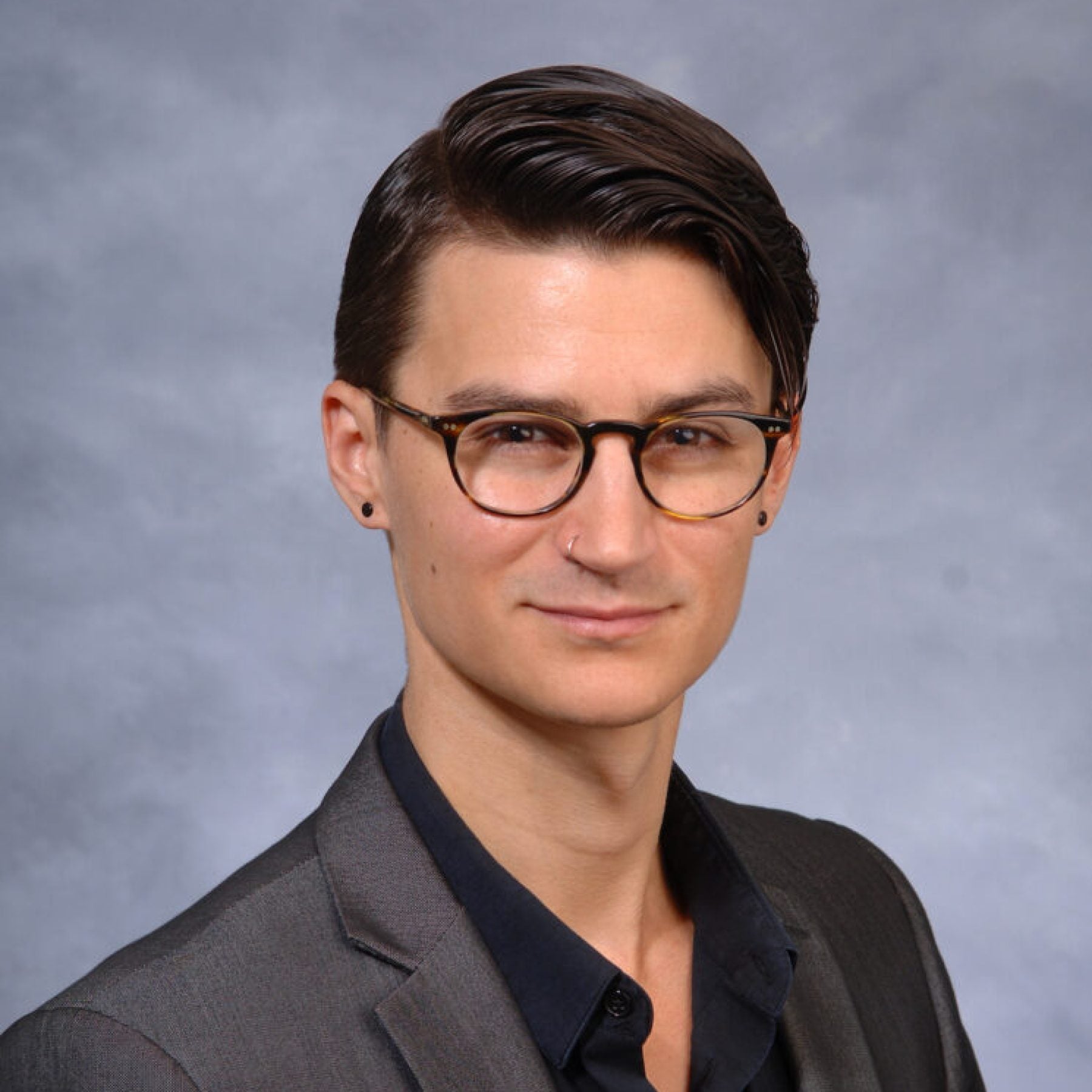 Joel Reynolds wears a gray blazer and black collared shirt, classes, and earring in a headshot against a gray background