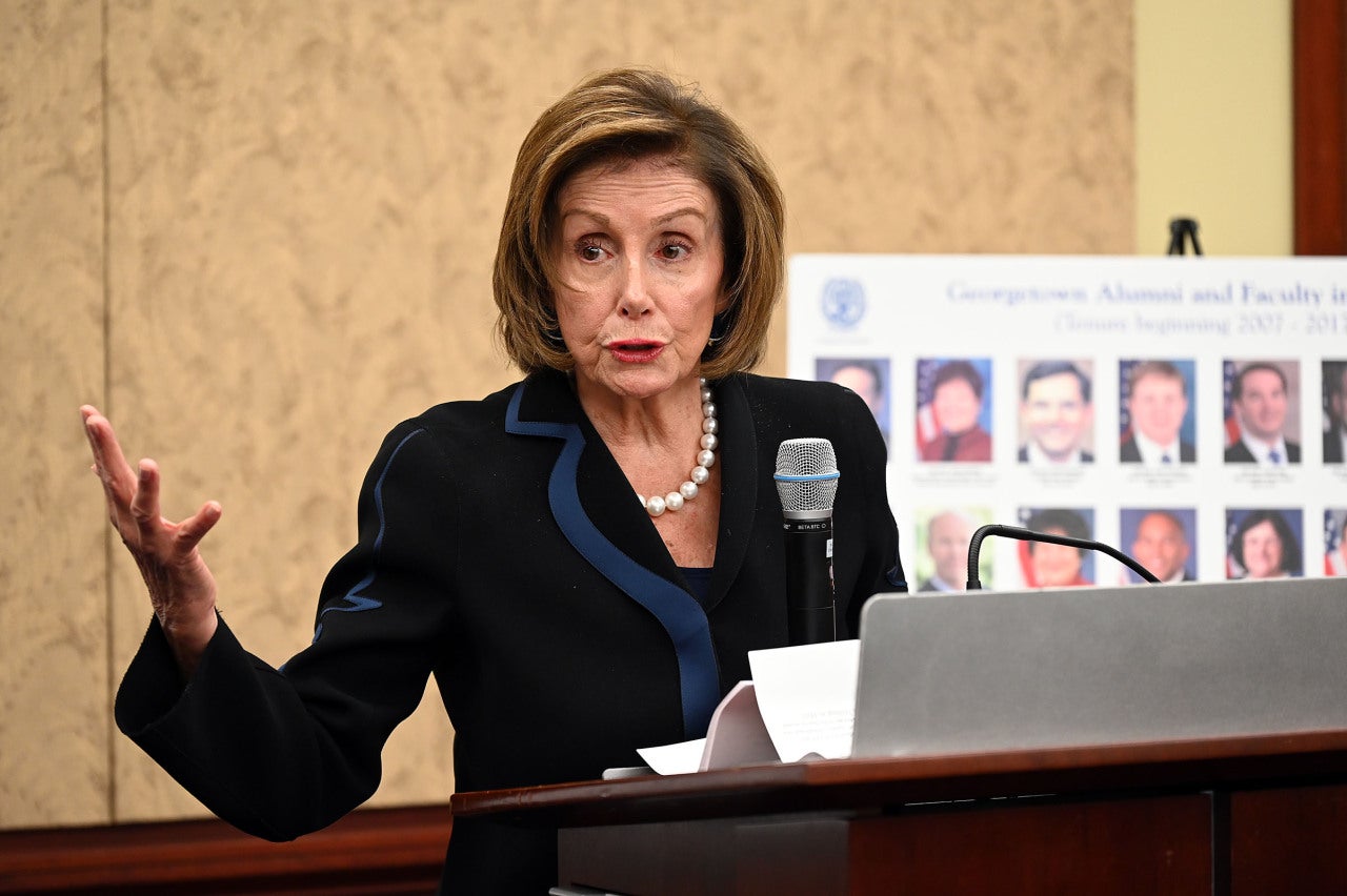 House Speaker Nancy Pelosi wears pearls and a blazer while speaking from behind a podium