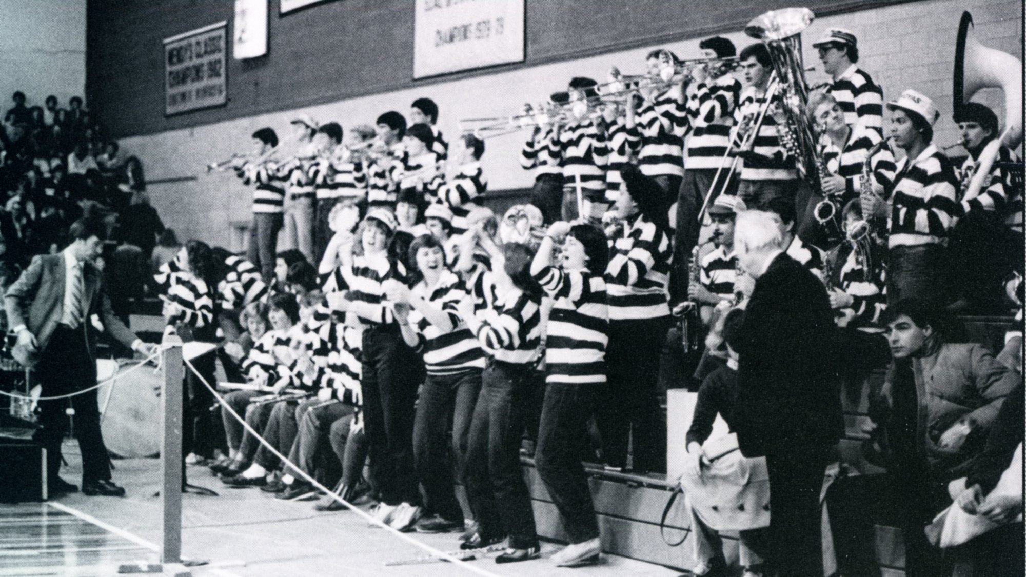 A black-and-white photo of the Pep Band, who are wearing striped long-sleeve shirts, standing on bleachers and cheering on a basketball game in 1984