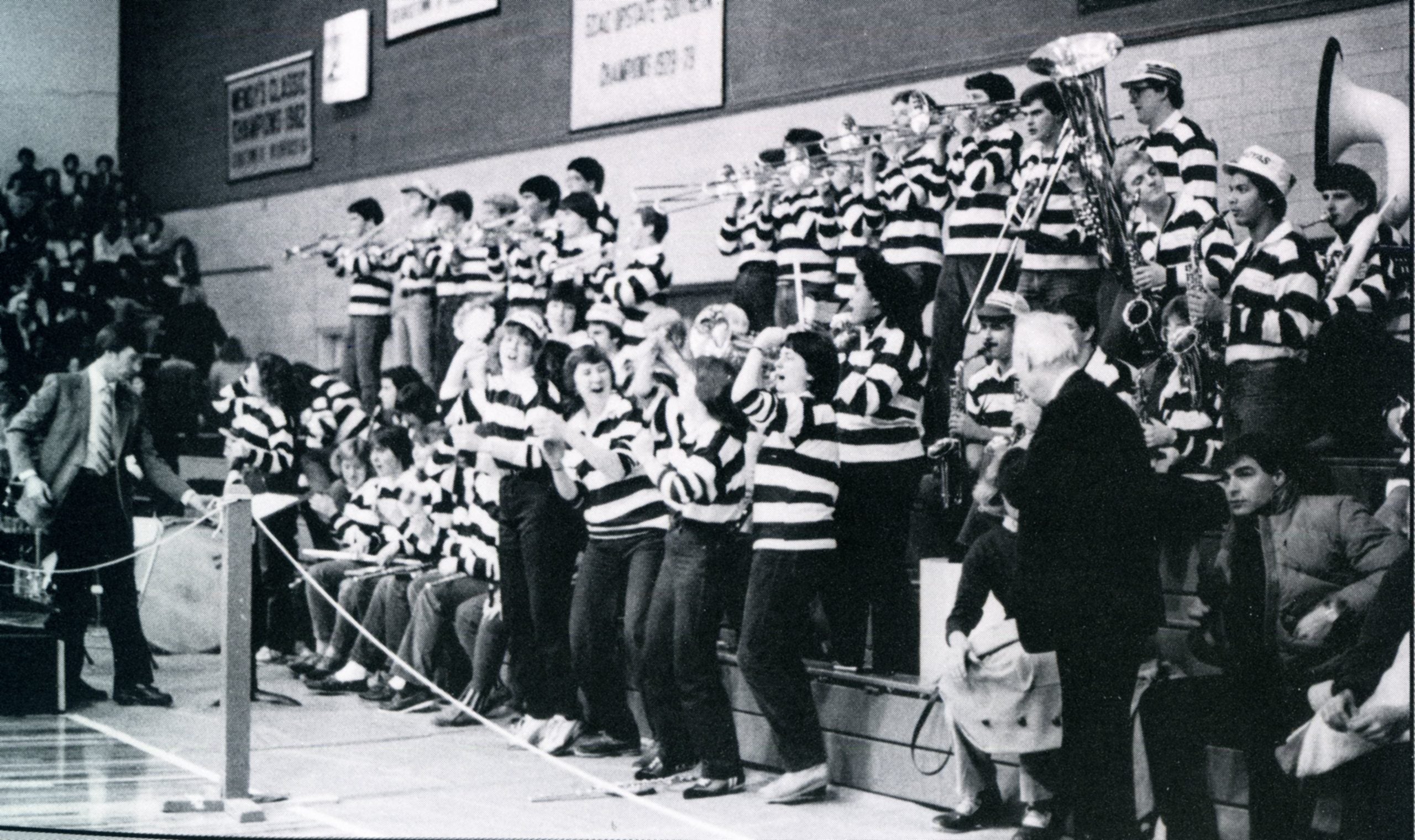 A black-and-white photo of the Pep Band, who are wearing striped long-sleeve shirts, standing on bleachers and cheering on a basketball game in 1984