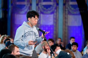 Young man wearing a gray Georgetown hoodie holds his phone while asking a question into a microphone