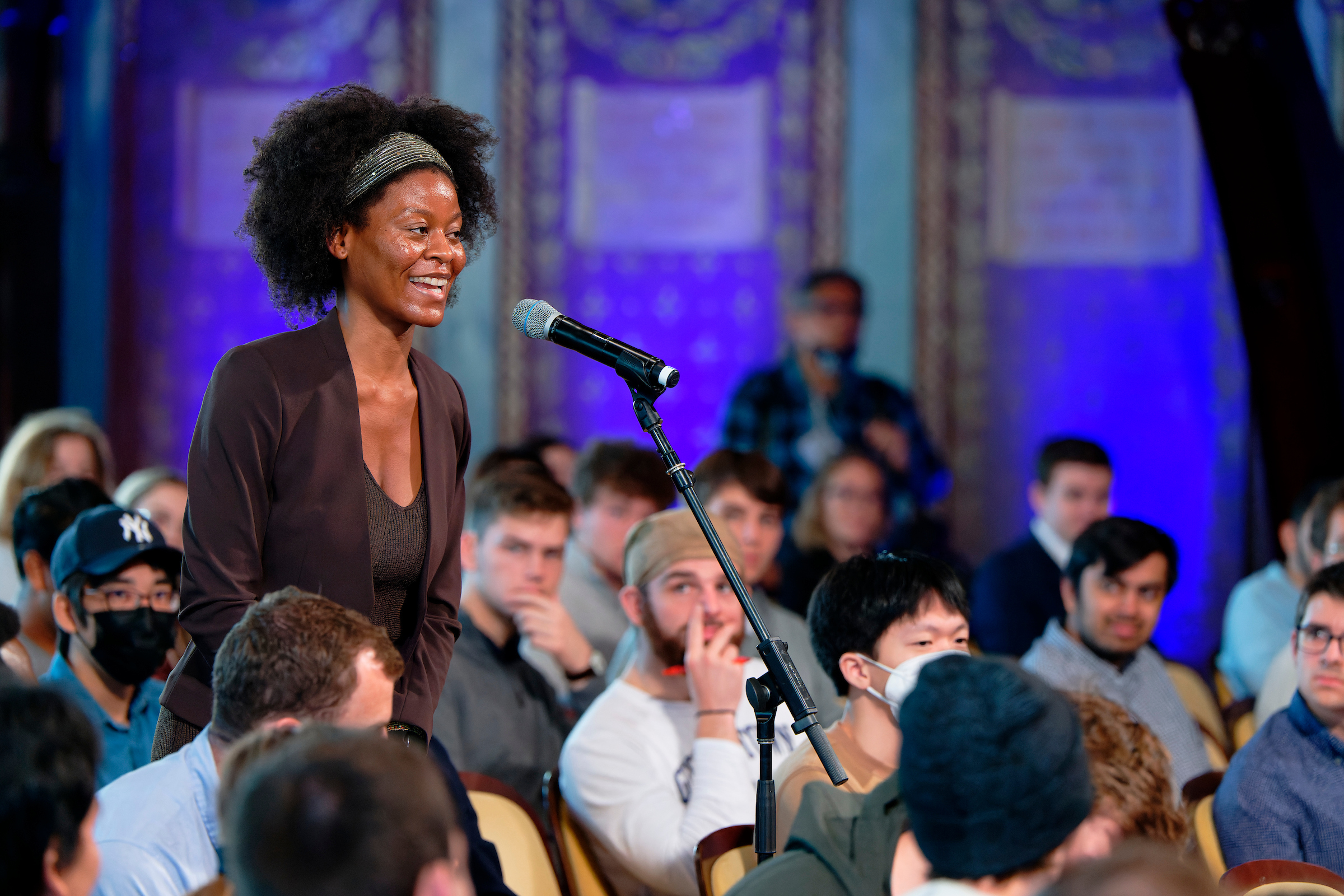 Young woman asks a question into a microphone while her peers look on from the audience