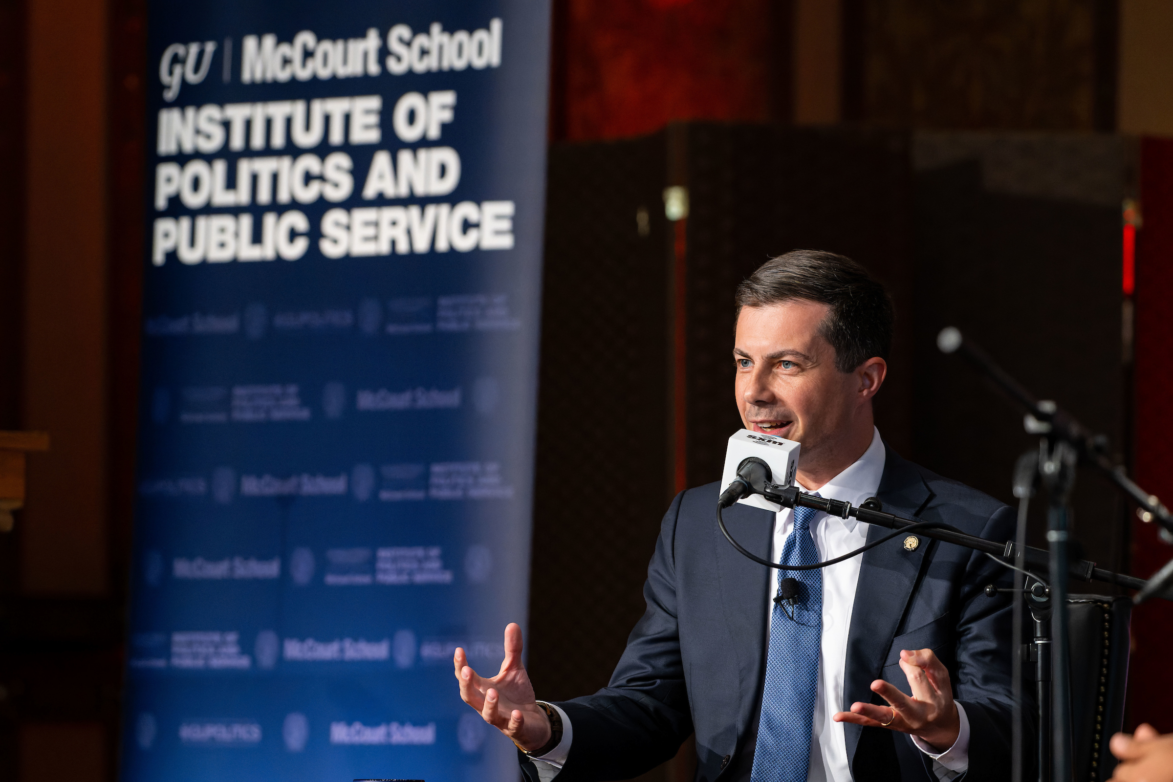 Pete Buttigieg talks into a microphone while seated onstage in front of a McCourt School Institute of Politics and Public Service sign