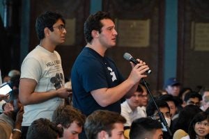 Young man holds a standing microphone as he asks a question and another young man waits in line behind him