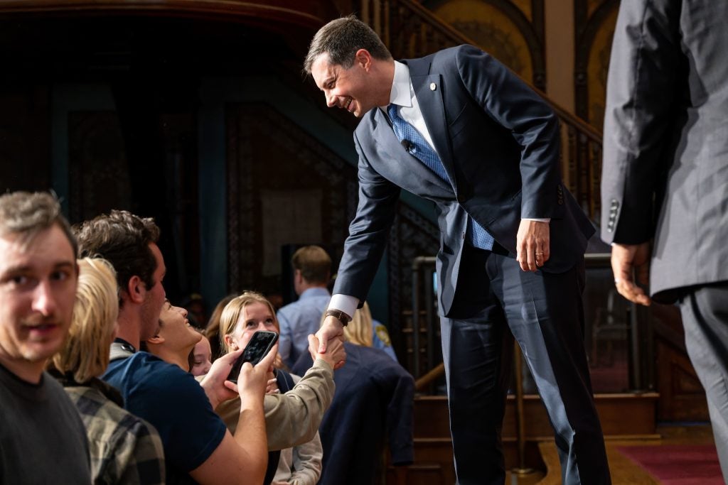 Pete Buttigieg wears a dark blue suit and shakes hands with the audience at the end of a stage