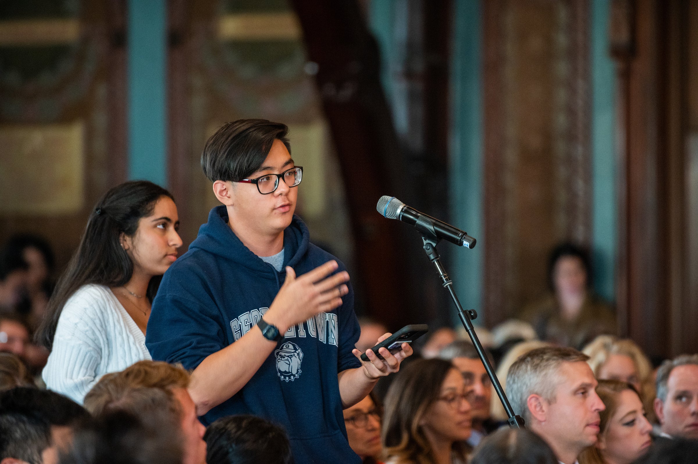 Young man wearing a dark blue Georgetown hoodie holds his phone while asking a question into a microphone