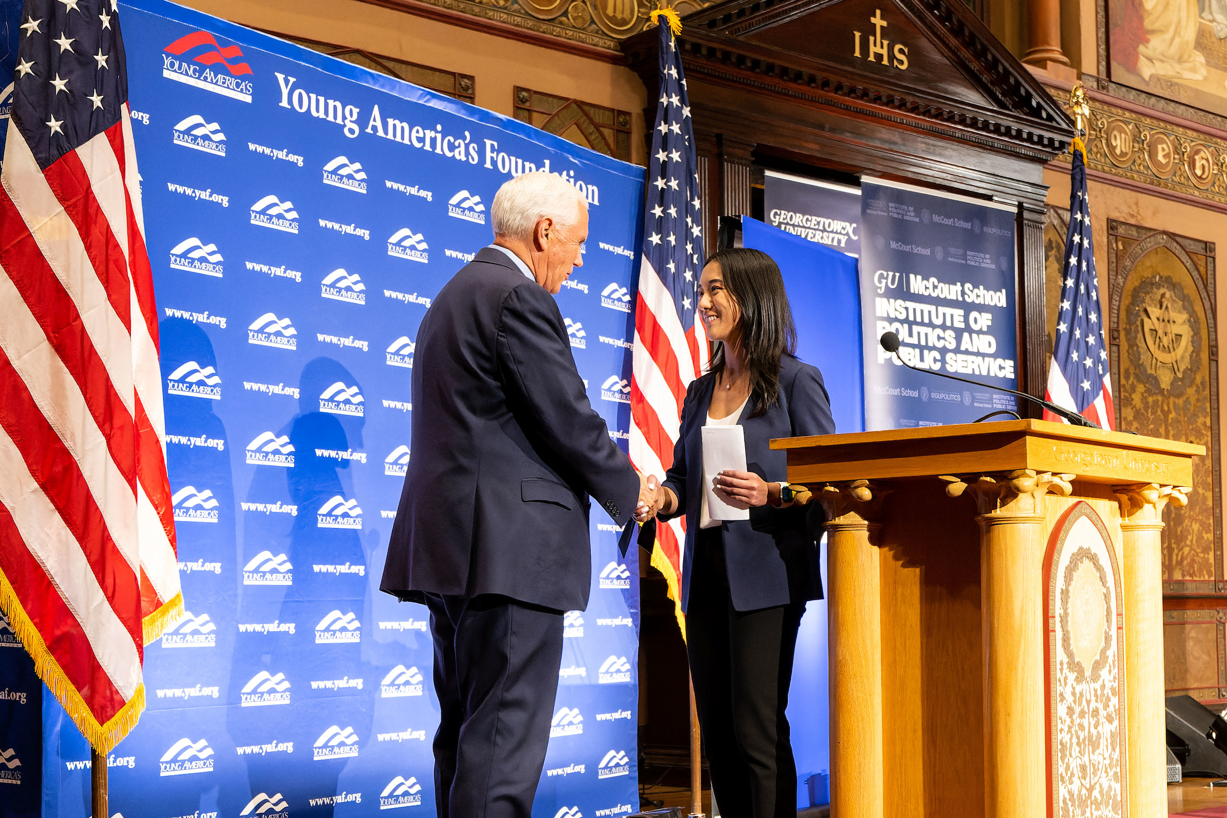 Mike Pence shakes the hand of a young woman behind a podium in front of a banner that says "Young America Foundation"