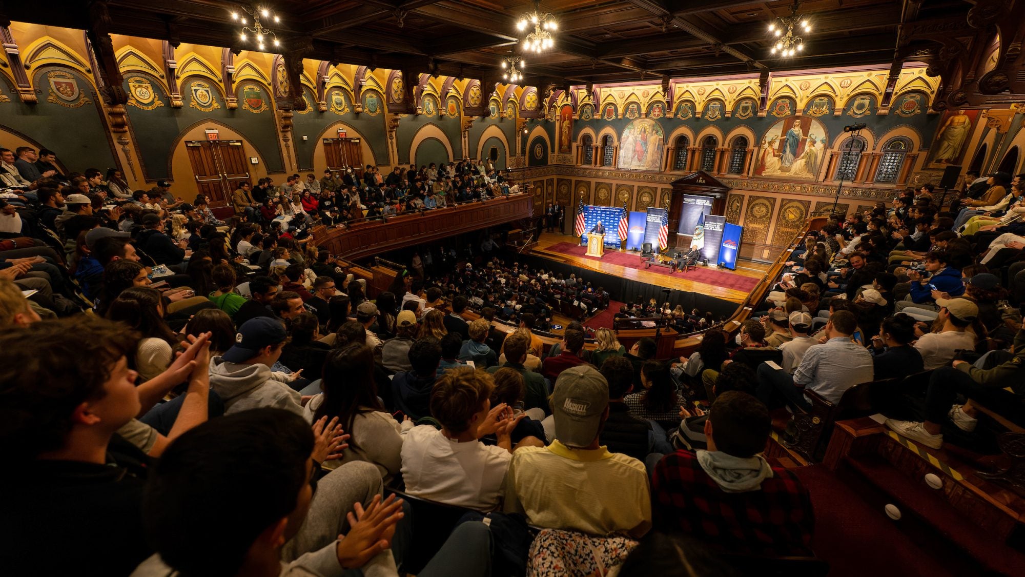 Large audience in an ornate auditorium looks at banners set up on a stage