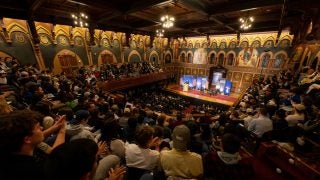 Large audience in an ornate auditorium looks at banners set up on a stage