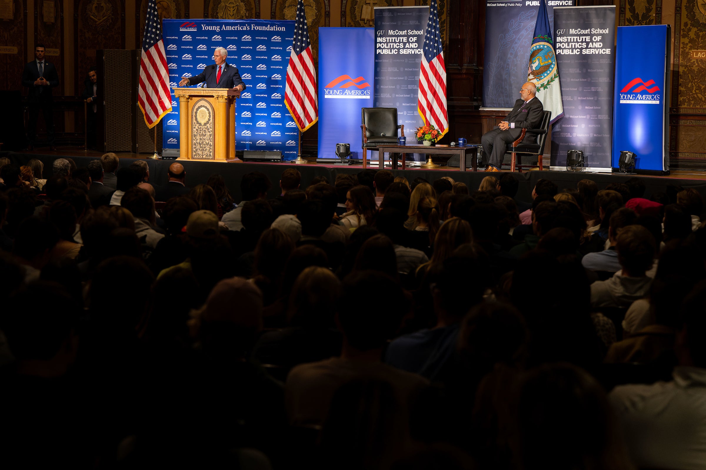 View from behind the audience watching Mike Pence speak from behind a podium while the moderator sits on a chair waiting to begin conversation