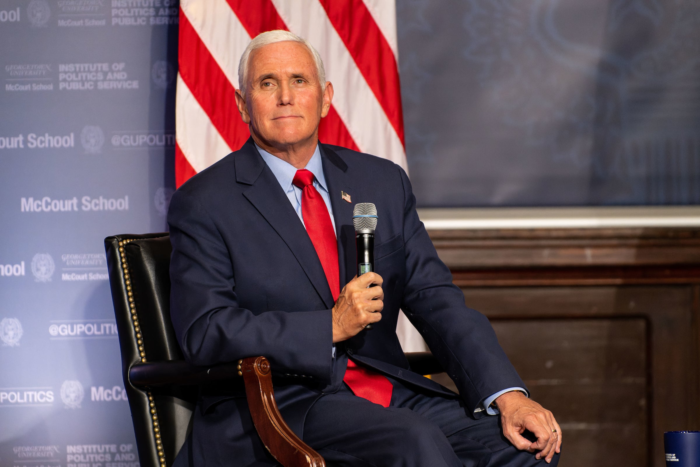 Mike Pence holds a microphone while wearing a dark blue suit and red tie while sitting onstage in front of an American flag