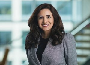  Feryal Faramarzi wears a gray blazer and black blouse in front of a staircase out of focus in the background