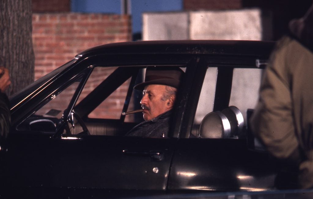 Actor Lee J. Cobb sits in a car while waiting for a scene of The Exorcist to film. He's smoking a cigar. 