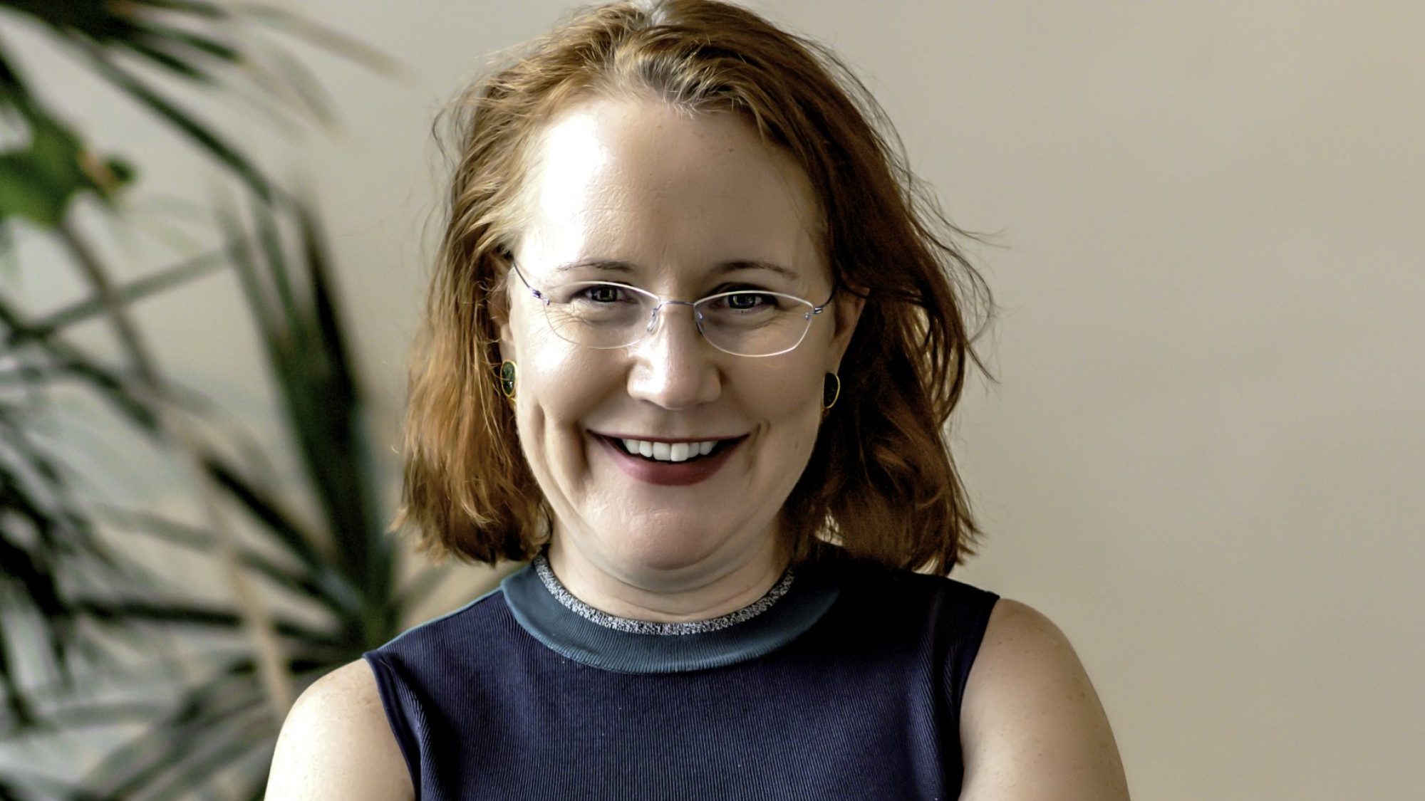 Woman with shoulder-length red hair and glasses smiles while folding her arms in front of a tropical potted plant