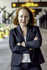 Woman with shoulder-length red hair and glasses wears a blazer and folds her arms with a hallway in the background