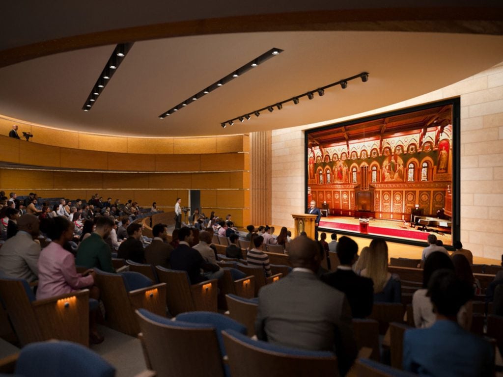 A rendering of the inside of an auditorium, with the lights dimmed and the backs of seated audience members facing a stage.