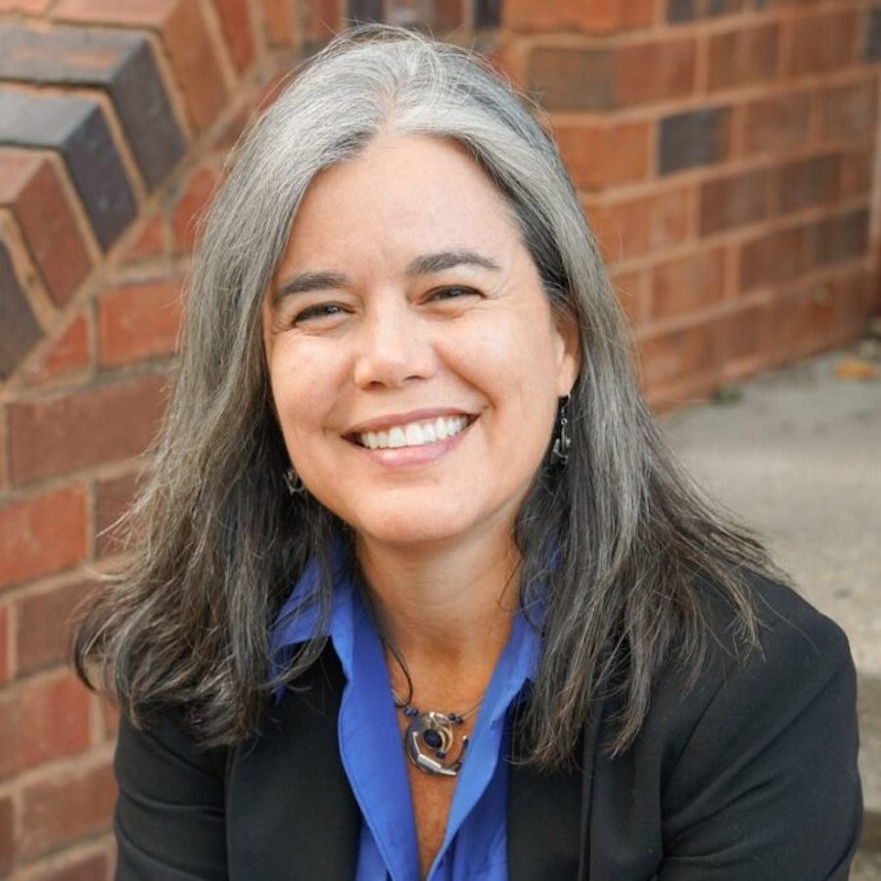 Roxanne Mirabal-Beltran has gray shoulder-length hair, a blue collared shirt and a black blazer in front of a brick railing