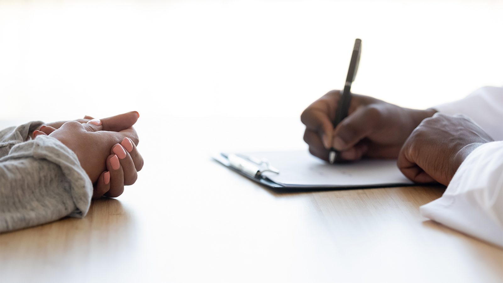 An image of two hands clasped across from a person writing on a notepad.