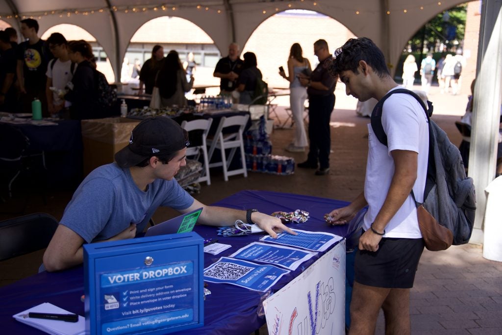 Alex Grass (SFS'22) sits behind a blue GU Votes table outside and points to instructions on how to register to vote as another student stands and looks on.