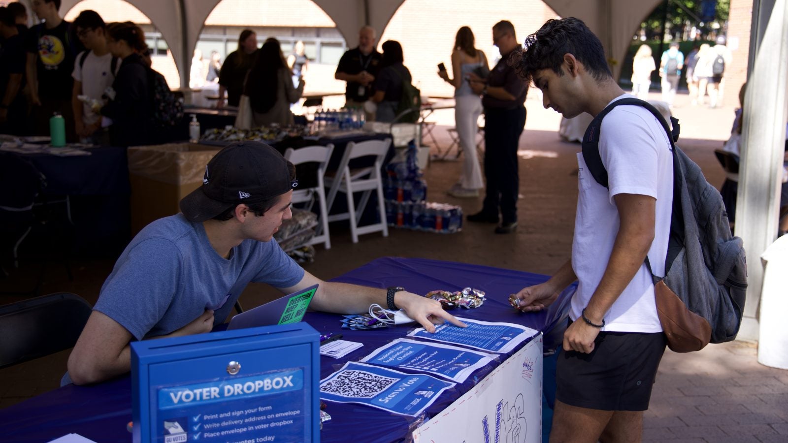 Alex Grass (SFS&#039;22) sits behind a blue GU Votes table outside and points to instructions on how to register to vote as another student stands and looks on.