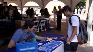 Alex Grass (SFS&#039;22) sits behind a blue GU Votes table outside and points to instructions on how to register to vote as another student stands and looks on.