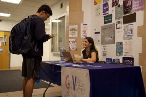 One stand stands in front of a table and another sits behind it answering questions about voter registration. The table is covered in a blue tablecloth and has a sign that says "GU Votes"