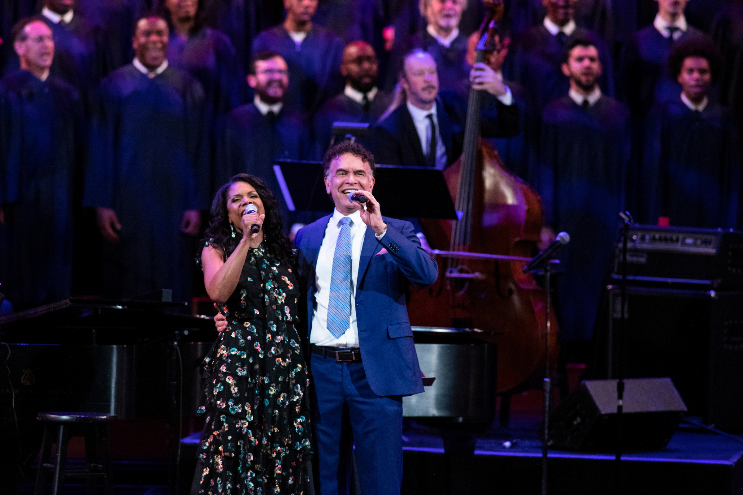 Audra McDonald and Brian Stokes Mitchell wrap their arms around each other while singing into mics out to an audience