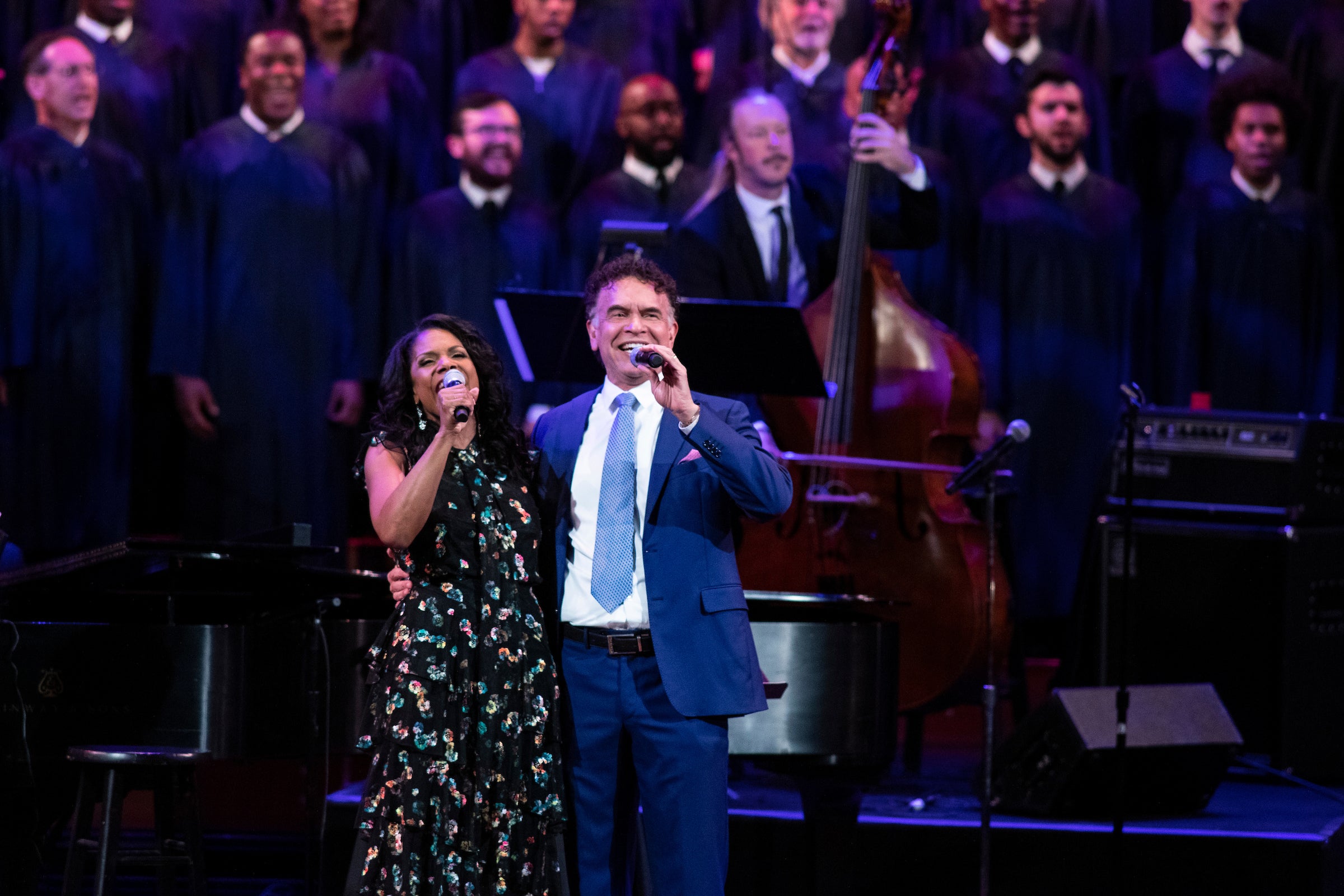 Audra McDonald and Brian Stokes Mitchell wrap their arms around each other while singing into mics out to an audience