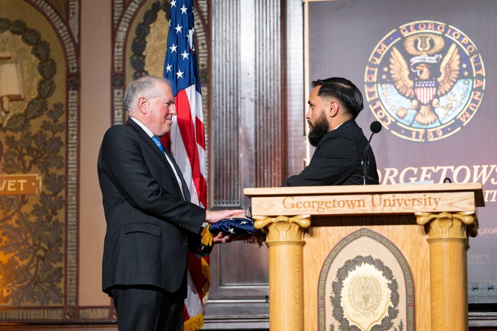 Frank Kendall receives a folded flag from Oswaldo Pazmino who stands behind a podium onstage in front of an American flag and Georgetown University sign