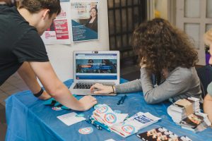Two students look at a laptop that says "Georgetown University" on National Voter Registration Day. One of the students is seated behind a blue table filled with voting resources and stickers while the other student stands to look at the screen.