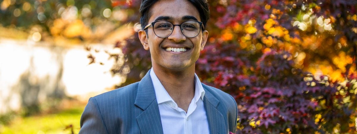 Atharv Gupta wears a white collared shirt and pinstripe gray blazer in front of a red, orange and yellow leaved tree