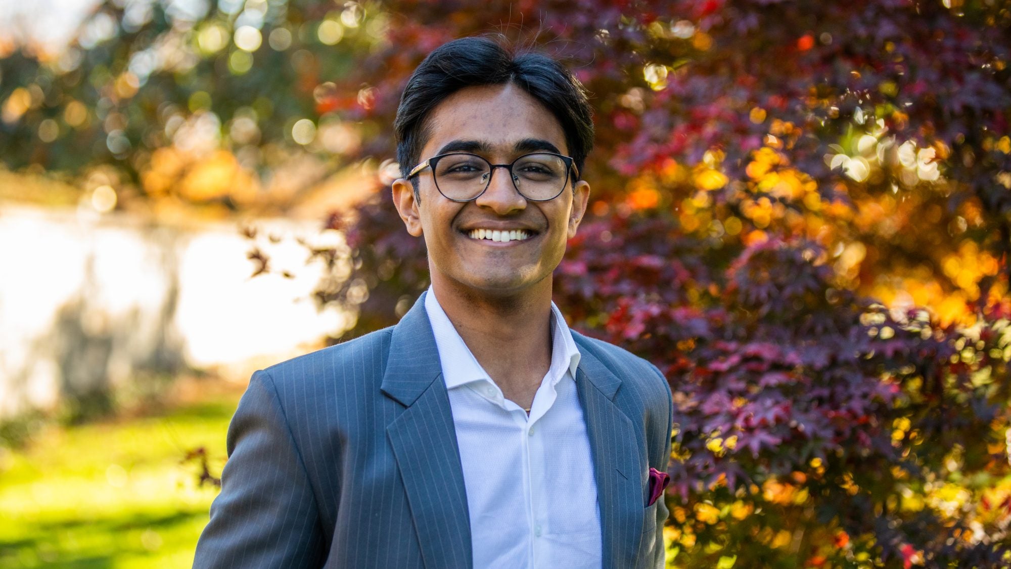 Atharv Gupta wears a white collared shirt and pinstripe gray blazer in front of a red, orange and yellow leaved tree