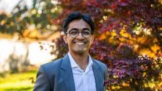 Atharv Gupta wears a white collared shirt and pinstripe gray blazer in front of a red, orange and yellow leaved tree