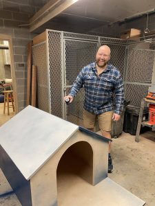 Ken Homan (G'25) smiles at the camera as he spray paints the roof of a wooden doghouse he is building for Georgetown's mascot. Ken is wearing a blue plaid button-down shirt, khaki shorts and blue tennis shoes.