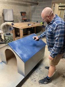 Ken Homan, a Jesuit brother, spray paints the roof blue of a new wooden doghouse he is building. He wears a plaid button-down shirt, khaki shorts and blue tennis shoes.