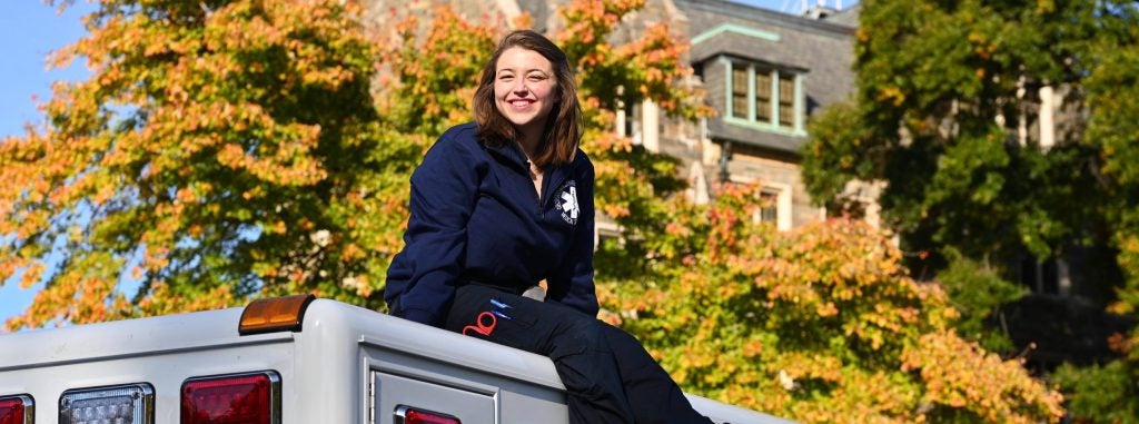 Isabella Turilli wears a blue jacket and pants while sitting on an ambulance in front of leavings changing from green to yellow