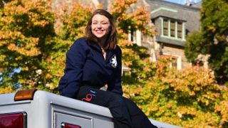 Isabella Turilli wears a blue jacket and pants while sitting on an ambulance in front of leavings changing from green to yellow