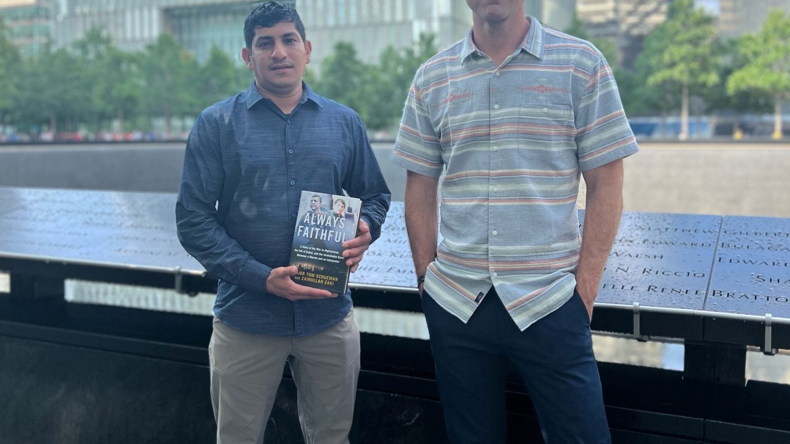 Two men stand next to each other in front of blue river and skyscrapers. The man on the left holds a book and is wearing a blue long-sleeved shirt and khakis. The man on the right wears a short-sleeve button-down and jeans.